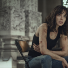A woman in her thirties with shoulder length brown hair, sits forward in a folding chair, clutching her side and with cuts and bruises peppering her arms. She looks concerned. She appears to be inside a chapel.