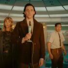 The cast of Loki stand facing the same direction with Tom Hiddleston center in a brown coat holding an old fashioned microphone