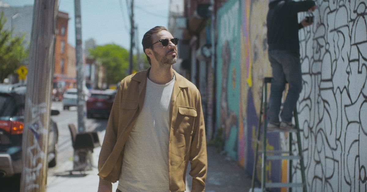 Nick Barili walks down a street with sunglasses and a tan jacket on in De La Calle