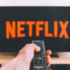 A TV with the Netflix logo sits behind a hand holding a remote