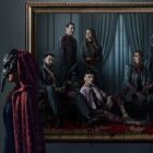 A promotional image for The Fall of the House of Usher showing a masked woman walk past a family portrait