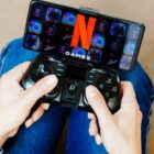 A person holding a gaming controller connected to a smartphone. The smartphone display shows the Netflix games logo