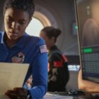 A woman with short black hair wearing a blue jumpsuit with an American flag patch on the shoulder is reading an important looking document. She looks concerned. In the background you see technical screens and some more space staff.