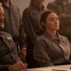 Sitting at a table are three people with very serious expressions. In the background there are more people standing, some with their arms crossed. They are all wearing the same gray jumpsuit.