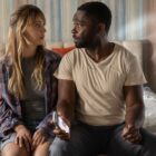 Kaley Cuoco as Emma and David Oyelowo as Dave sit together on a bed looking shocked as David holds a phone in Role Play.