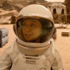 Close up of a female astronaut on Mars. In the background there are several containers and a white building.