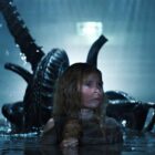 A young girl with long blonde hair is wading in water up to her armpits. She looks scared. Just behind her in the water is a dark, smooth-headed scary-looking alien with a sharp, whip-like tail. The alien (xenomorph) is getting ready to pounce on the girl.