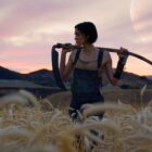 Kora stands in a field of wheat with a scythe over her shoulders in Rebel Moon Part 1