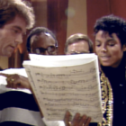 Two white men and two Black men gather around sheet music inside a recording studio.