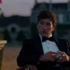 A still from the movie Saltburn of Barry Keoghan as Oliver Quick in a fancy tuxedo sat outside in a chair with a huge looming stately home in the background.