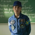 A woman wearing a blue astronaut jumpsuit decora ted with several different patchers, including a Swedish flag. She is also wearing a blue baseball cap. She is standing in front of a black board with some chalk writing on.