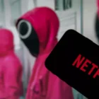 Netflix logo on the display of a smartphone in front of a television with the new series "Squid Game" ( focus on Netflix logo )
