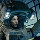 a woman in a bulky tan spacesuit looks at something on a metallic wall