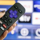 A hand holds a Roku remote in front of a TV with the Roku home screen.