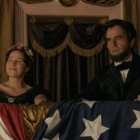 Actors playing President Lincoln and his wife sit in a theater box, an American flag draped in front of them.