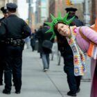 Elsbeth Tascioni (Carrie Preston), poses in a novelty Statue of Liberty hat behind NYPD officers while looking to camera in a promo image for CBS