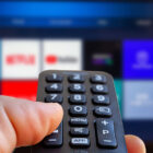 A man is holding a remote control of a smart TV in his hand. In the background you can see the television screen with streaming entertainment apps for video on demand