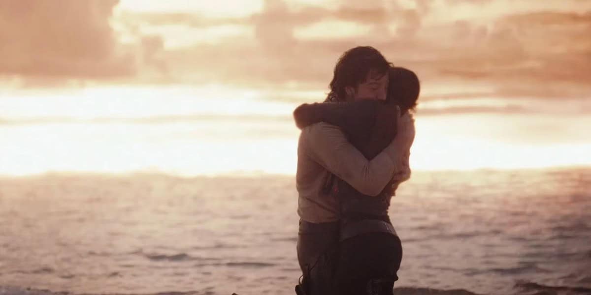 Two people embrace each other tightly on a beach at sunset, with the ocean and a colorful sky of orange and yellow hues in the background.