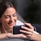 A woman with long brown hair smiles while looking at her smartphone. She is wearing a white shirt and has white earphones in her ears. The background is blurred, focusing attention on her joyful expression.