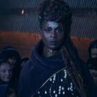 characters in robes look at one another in a dingy cave-like structure