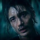 A person with wet hair and a mustache looks intently at the camera in a dimly lit setting. Their expression is serious, and their face glistens with moisture, suggesting a tense or dramatic scene.