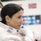 an astronaut with long brown hair stands in a bulky white spacesuit with the helmet off inside of a training facility with white walls