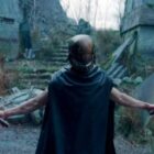 Qimir/The Stranger in a dark cape stands in a post-apocalyptic environment, holding two red lightsabers reminiscent of Star Wars. The surroundings include ruined buildings, overgrown vegetation, and debris. The figure has a menacing stance with arms extended, lightsabers ignited.