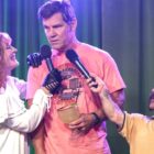 An unrecognizable Glenn Close wearing white in profile on stage with Josh Brolin in a pink shirt holding a big margarita and Peter Dinklage with a big mustache holding up a mic in Brothers