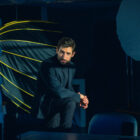 A man with a beard and short dark hair, wearing a black turtleneck and blazer, sits in a dimly lit photography studio. He is illuminated by moody lighting, with a serious and contemplative expression.