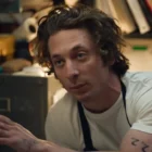 Person talking in a cluttered room, wearing a white shirt.