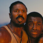 Smoke and Sammie holding each other as they watch a fire burn off-camera in Sinners