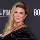 Actress Gina Carano stands in front of a black backdrop. She smiles into the camera and wears a black dress with long blonde hair draping over her chest.