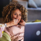 A woman eating popcorn on a sofa watching her laptop