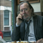 A man (Gary Oldman playing the character of Jackson Lamb) sitting at a table by the window in a cafe. He has a full English breakfast on a plate in front of him and is holding a mobile phone to his right ear.