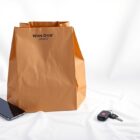 A minimalist studio still life featuring a stack of paper grocery bags, a smartphone, and a set of car keys arranged elegantly on a clean, white background, conceptually representing the convenience and on-demand nature of the new Winn-Dixie grocery delivery service.