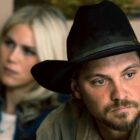 Kayce looks off into the distance while a blonde woman is sat behind him