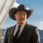 John Dutton, played by Kevin Coster, stands in his cowboy hat and suit with Montana mountains in the background.