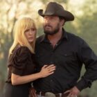 A blonde woman stands close to a man with a dark brown beard in a cowboy hat and puts her hand on his stomach.
