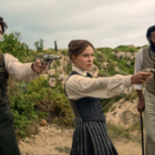 A woman in Victorian period costume stands in the desert pointing a small revolver. To her right is a white man doing the same. To her left, a man of Indian descent leaning on a walking stick.