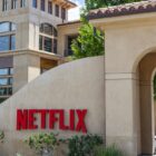 A wide, sunlit exterior shot of the Netflix corporate campus. In the foreground, a curved, light beige stucco wall prominently displays the red, sans-serif 'NETFLIX' logo. Behind the wall, a modern building with multiple windows and balconies is visible. To the right, an arched entryway leads to another part of the campus. Green trees and shrubs are scattered around the base of the wall and buildings under a bright blue sky.