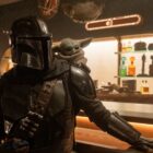 Mandalorian wearing a helmet, standing at a bar, with Grogu perched on shoulder.
