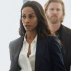 Joe walks along a hallway with a man behind her