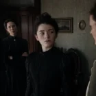 Ella Beatty as Lizzie Borden stands in a dark period scene holding an axe.